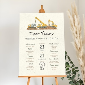 May include: A decorative sign on an easel with the text "Two Years Under Construction." The sign includes a watercolor illustration of construction vehicles and lists the child's nickname, weight (23 pounds), height (21 inches), favorite foods, toys, book, and song.