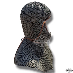 Puede incluir: Se muestra un coif de cota de malla, un tipo de cobertura para la cabeza medieval. El coif está construido con numerosos anillos metálicos pequeños e interconectados. Se ve una correa de cuero. El logotipo de The Artisan Store está impreso en el coif.