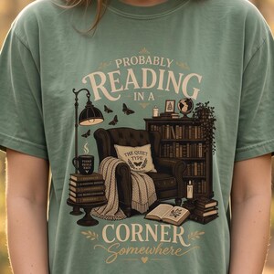 May include: A sage green short-sleeve t-shirt featuring a graphic illustration of a cozy reading nook with an armchair, a bookshelf, a lamp, and stacks of books. The text reads: PROBABLY READING IN A CORNER SOMEWHERE and THE QUIET TYPE.