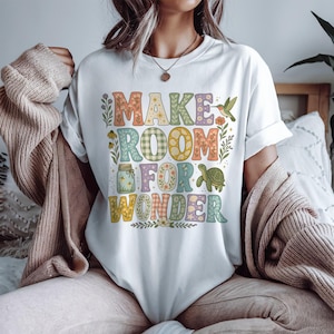 May include: A white short-sleeve t-shirt featuring the text "MAKE ROOM FOR WONDER" in large, multi-colored block letters. The design includes floral patterns, a mason jar, a hummingbird, and a turtle, all rendered in soft, muted earth tones.
