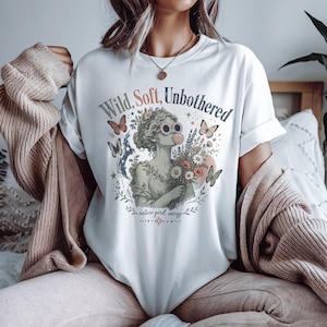 May include: A white t-shirt featuring a graphic of a classical statue wearing sunglasses and blowing a bubble, surrounded by flowers, butterflies, and stars. The text reads Wild, Soft, Unbothered and nature girl energy in a decorative font.