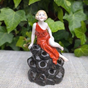 May include: A porcelain figurine of a person with blonde hair wearing a bright red dress, seated atop a dark, textured, rock-like base with multiple circular openings. The figure has pale skin and is positioned in a relaxed, seated pose.