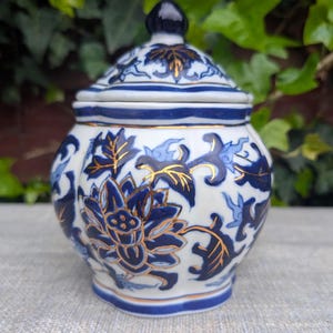 Puede incluir: Un pequeño tarro de cerámica con tapa, adornado con un diseño floral azul y blanco y detalles dorados. El tarro tiene un cuerpo redondeado y una tapa a juego con un remate azul oscuro. El patrón floral incluye flores de loto y hojas.