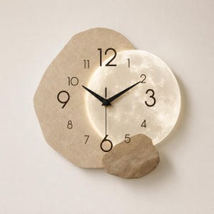 May include: A wall clock featuring an irregular beige textured backing and a circular, illuminated moon-like face. The clock has black hour and minute hands, a thin second hand, and black numerals from 1 to 12 printed on the surface.