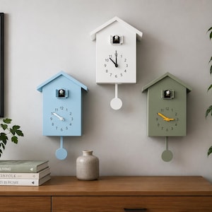 May include: Three house-shaped wall clocks in light blue, white, and olive green. Each clock features a small birdhouse opening at the top, a circular clock face with black numbers, two hands, and a matching circular pendulum hanging from the bottom.