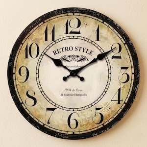 Könnte beinhalten: Eine runde Wanduhr mit einem verwitterten cremefarbenen Zifferblatt, schwarzen arabischen Ziffern und einem schwarzen Rahmen. In der Mitte steht der Text RETRO STYLE, 1904 de Paris und 21 boulevard Batignolls. Zwei schwarze Zeiger zeigen die Zeit.