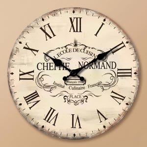 Può includere: Un orologio da parete rotondo con quadrante color crema, numeri romani neri e lancette in metallo nero. Al centro è riportato il testo L'ECOLE DE CUISINE, CHEFIE NORMAND, Institut Culinaire Français e l'anno 1930 in caratteri decorativi.