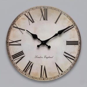 May include: A round wall clock with a distressed off-white face featuring black Roman numerals and black ornate hands. The centre of the clock face displays the text London, England in a black script font. The design includes a thin black border.