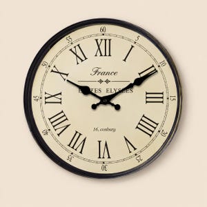 May include: A round wall clock with a black frame and a cream-coloured face featuring black Roman numerals. The dial displays the text France, CHAMPS ELYSEES, and 16, conburg. It has two black ornate hands and a minute track with Arabic numerals.