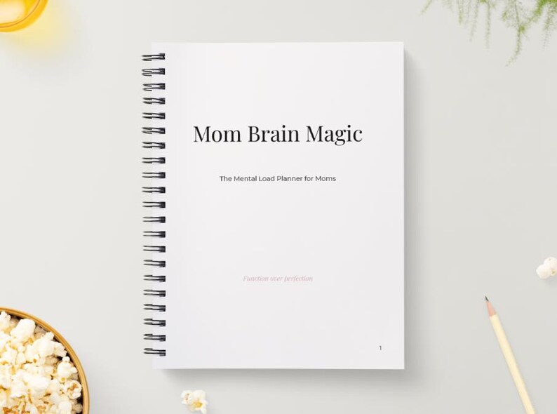 May include: A white spiral-bound notebook titled "Mom Brain Magic" with the subtitle "The Mental Load Planner for Moms." The cover also includes the phrase "Release your perfection." A pencil and a bowl of popcorn are in the background.