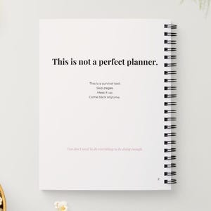 May include: A white spiral-bound planner with the text "This is not a perfect planner." Additional text includes "This is a survival tool. Skip pages. Mess it up. Come back anytime."