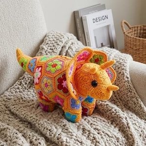 Könnte beinhalten: Ein handgefertigtes, gehäkeltes Triceratops-Dinosaurierspielzeug in Orange-, Gelb-, Grün-, Rosa- und Blautönen. Der Dinosaurier hat ein detailliertes Blumenmuster und ist auf einer grauen Decke drapiert. Das Spielzeug hat schwarze Knopfaugen und drei Hörner.
