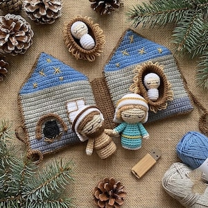 Puede incluir: Escena de nacimiento tejida a ganchillo con una casa azul y gris, detalles de estrellas y pequeñas figuras. Incluye un bebé en un pesebre y otras dos figuras. Sobre una superficie de arpillera con piñas y vegetación.