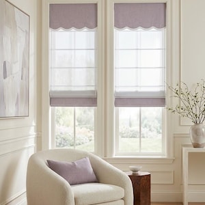 May include: Two windows featuring light purple fabric roman shades with a scalloped bottom edge and white trim. The shades are layered over sheer white panels. A cream-colored textured armchair with a matching purple rectangular pillow sits below.
