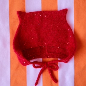 May include: A bright red knitted bonnet featuring two pointed ear-like projections at the top. The fabric is embellished with small, scattered red sequins throughout. The bonnet includes two knitted chin straps tied in a bow at the bottom.