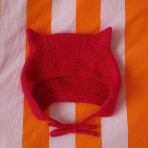 May include: A bright red knitted baby hat with pointed ears and tie strings. The hat is made of a soft, textured yarn and is set against a striped orange and white background. The hat's design resembles a cat or animal.