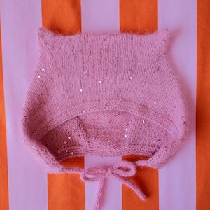 May include: A soft pink knitted bonnet featuring two pointed cat-like ears at the top. The fabric has a fuzzy texture with scattered small silver sequins throughout. The bonnet includes a matching knitted chin strap tied in a bow.