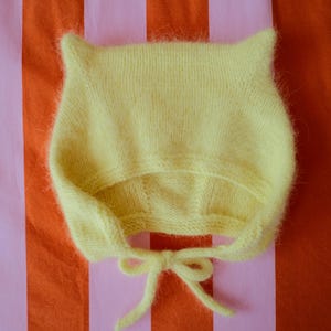 May include: A pale yellow knit bonnet featuring two pointed cat-like ears at the top and a soft, fuzzy texture. The bonnet includes two thin knitted ties that are knotted together at the bottom to secure the head covering in place.