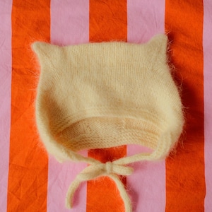 May include: A light yellow knitted baby bonnet with ear-like points and a tie closure. The bonnet is made of a soft, fuzzy yarn. The background is a striped pattern of orange and pink.