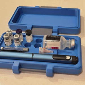 Könnte beinhalten: Ein blauer Kunststoffkoffer mit einem blauen Injektionsstift, einer durchsichtigen Glasampulle mit schwarz-weißem Etikett, vier kleinen Glasampullen mit farbigen Verschlüssen, einer durchsichtigen Nadelkappe und einer Nadel in einer Hülle.