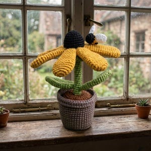 Puede incluir: Una flor de ganchillo hecha a mano con pétalos amarillos, un centro negro y un tallo y hojas verdes. Una pequeña abeja de ganchillo descansa sobre un pétalo. La flor está en una maceta de ganchillo gris.