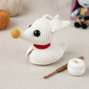 May include: A crocheted Zero dog from The Nightmare Before Christmas. The white dog has black eyes, a red collar, and a yellow nose. A wooden crochet hook and a ball of white yarn are next to the dog.