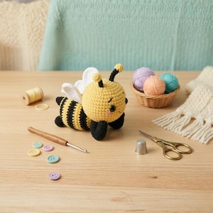 May include: A handmade crochet bee with yellow, black, and white details. The bee is lying on a wooden surface, with a crochet hook, buttons, yarn, and scissors nearby. The bee measures approximately 10 cm long.