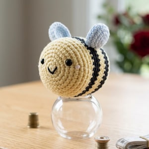 May include: A handmade crochet bee with a smiling face, black stripes, and light blue ears. The bee is perched on a small, clear glass jar. The bee is yellow and black, and the jar is sitting on a wooden surface.