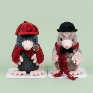 May include: Two crocheted mole figures, one in a red cap, scarf, and magnifying glass, the other in a black bowler hat, scarf, and bow tie. The moles are gray, tan, and cream-colored, standing on white platforms against a light green background.