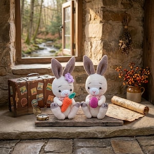 Puede incluir: Dos conejos de crochet hechos a mano, uno con una zanahoria y el otro con un huevo. Son blancos con detalles rosas y morados, sentados en una tabla de madera. Una maleta vintage y una brújula están cerca.