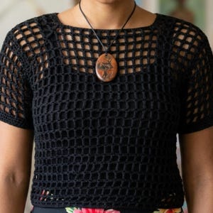 Puede incluir: Un top negro de ganchillo con cuello cuadrado y mangas cortas. El top tiene un diseño abierto, similar a una red. Un colgante marrón y naranja cuelga de un cordón negro alrededor del cuello. El top se lleva sobre una prenda con estampado floral.