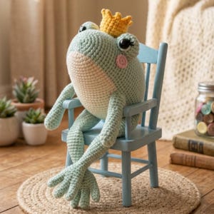 May include: A crocheted frog doll in shades of light blue and cream, wearing a yellow crown, sits on a small blue chair. The frog has black eyes and pink cheeks. The doll is placed on a round, woven mat.