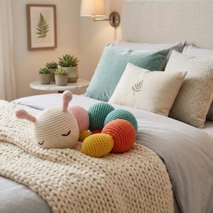 May include: A collection of colorful, crocheted toys and pillows on a bed. The toys include a cream-colored creature with pink ears and closed eyes, and several round balls in teal, orange, pink, and yellow. The bed is covered with a cream-colored knitted blanket and several pillows.