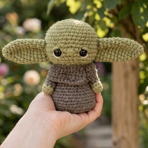 May include: A handmade crochet doll in the likeness of a popular character. The doll features large, pointed ears, big black eyes, and is dressed in a brown robe. The doll is held in a hand against a blurred green background.
