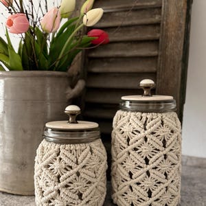 Può includere: Due barattoli di vetro con coperture in macramè. I barattoli hanno coperchi in legno con pomelli in ottone. Il macramè è color crema e i barattoli sono di diverse dimensioni. Un vaso di tulipani è sullo sfondo.