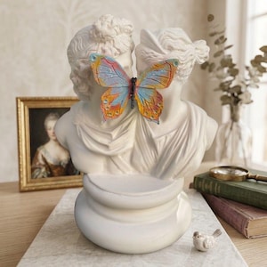 Könnte beinhalten: Eine weiße Skulptur von zwei klassischen Büsten, die sich gegenüberstehen, mit einem bunten Schmetterling dazwischen. Die Skulptur steht auf einem Marmorsockel, mit einer kleinen Vogelfigur in der Nähe. Ein gerahmtes Porträt und Bücher sind im Hintergrund.