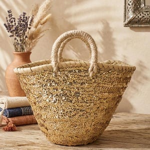 Puede incluir: Un bolso tote tejido con un exterior de lentejuelas. El bolso tiene una forma redondeada con un borde trenzado y asas de cuerda. El bolso está sobre una superficie de madera, con un jarrón de flores secas y una pila de textiles doblados en el fondo.