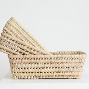 May include: Two woven rectangular baskets. The larger basket is light beige and has a rectangular shape. The smaller basket is placed on top, angled, and has a similar woven design. The baskets are made of natural fibres.