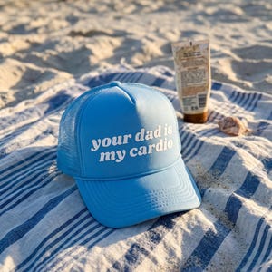May include: A light blue trucker cap with a mesh back and a foam front panel featuring the white text "your dad is my cardio" printed in a bold, rounded font. The cap has a matching blue brim and a decorative cord across the base of the crown.