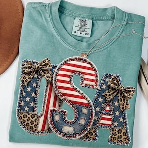 Könnte beinhalten: Ein kurzärmeliges T-Shirt in Blaugrün mit einem Comfort Colors-Etikett. Das Shirt zeigt eine große, dreidimensionale Applikation der Buchstaben USA, verziert mit rot-weißen Streifen, blauen Sternenmustern und Leopardenmuster-Akzenten mit kleinen Schleifen.
