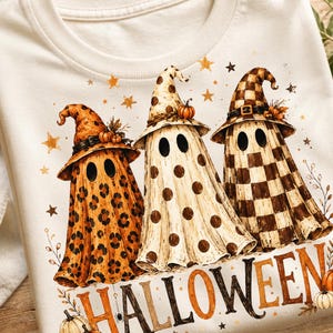 May include: A cream-colored t-shirt featuring a graphic of three ghosts wearing witch hats with leopard print, polka dot, and checkered patterns. The word HALLOWEEN is printed in orange and black letters below, surrounded by small pumpkins and stars.