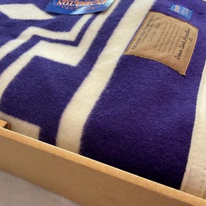 Puede incluir: Una manta de lana doblada con un patrón geométrico en zigzag de color morado y crema. Incluye una etiqueta de la marca Pendleton azul, una etiqueta tejida azul y un parche rectangular color canela con el texto "Onyayenaka Niamayou" y "Deer Dark Mountain 52".