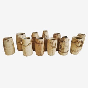 May include: A collection of weathered, cylindrical wooden vases in varying heights and widths. Some vases have rope handles or decorative rope accents. The vases have a natural, aged appearance with visible wood grain and a light brown color.