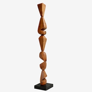 May include: A tall, abstract wooden sculpture with a series of stacked, geometric shapes. The sculpture is made of light brown wood and stands on a small, square black base. The shapes include cones, diamonds, and rounded forms.