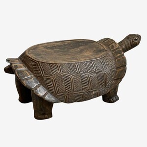 May include: A dark brown, carved wooden turtle sculpture with a flat top, likely a stool or small table. The turtle's shell features detailed geometric patterns. The head and legs are extended, giving the piece a lifelike appearance.
