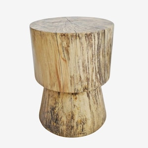 May include: A light-colored wooden stool with a cylindrical top and a wider, conical base. The wood grain is visible, with natural cracks and variations in color. The stool is approximately 18 inches tall.