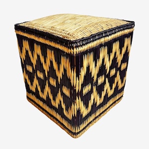 May include: A square woven storage ottoman with a geometric pattern. The ottoman features a natural tan top and a black and tan woven design on the sides. The edges are trimmed in black.