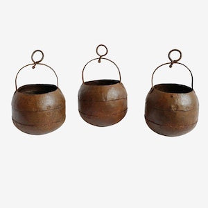 May include: Three round, dark brown metal pots with a weathered, oxidized finish. Each pot features a horizontal band around the center, visible rivets, and a thin, arched metal handle with a circular loop at the top for hanging or carrying.