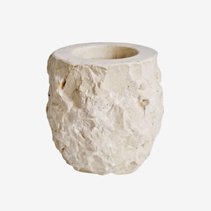 May include: A cream-colored, stone-like candle holder with a rough, textured exterior. The cylindrical holder has a wide opening at the top and a solid base. The natural, organic design adds a rustic touch to home decor.