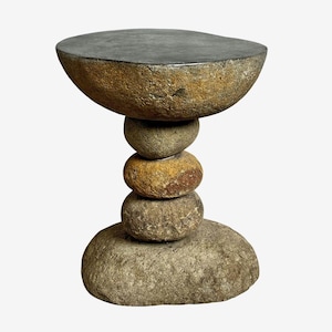 May include: A unique, handcrafted side table made of natural stones. The table features a dark gray, flat top supported by a stack of rounded stones in shades of brown and gray. The base is a large, flat stone.